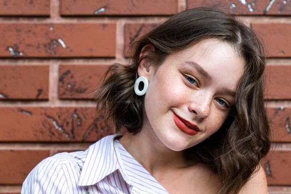 붉은 입술, 주근깨, 파란 눈으로 붉은 벽돌 벽에 고립된 카메라를 보며 웃고 있는 행복 한 갈색 머리 소녀의 근접 사진 — 스톡 사진