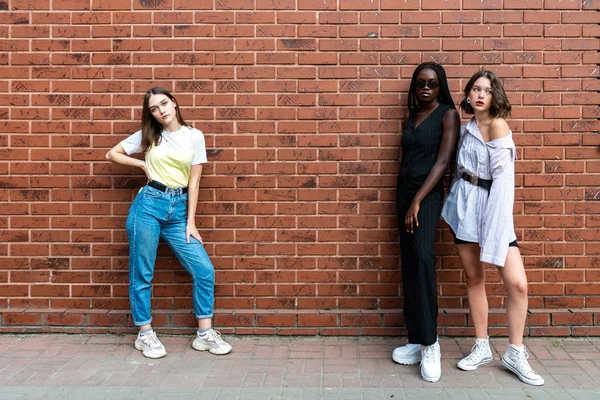 Foto multi étnica, dos chicas europeas y una afroamericana en ropa casual de pie en lados opuestos aislados sobre la pared de ladrillo rojo — Foto de Stock