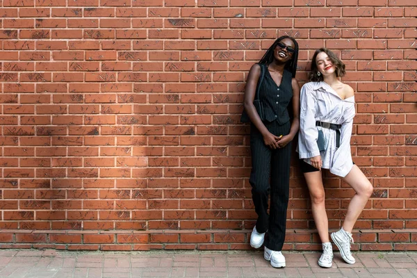 Glad leende multietniska flickor i casual kläder står tillsammans och tittar på kameran isolerad över röd tegelvägg — Stockfoto