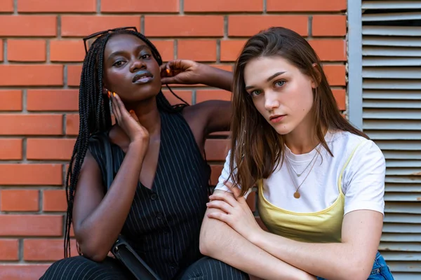 Cintura hacia arriba retrato de joven morena europea en ropa casual agachada junto con chica afroamericana en ropa de rayas negras aisladas sobre la pared de ladrillo rojo —  Fotos de Stock