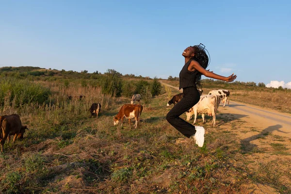 Młoda uśmiechnięta afrykańska dziewczyna skacze po polu, krowy pasą się na tle — Zdjęcie stockowe