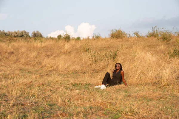 黒い服の若いアフリカの女の子は、夏の晴れた日を楽しんで、乾燥した芝生のフィールドの間に座っている — ストック写真