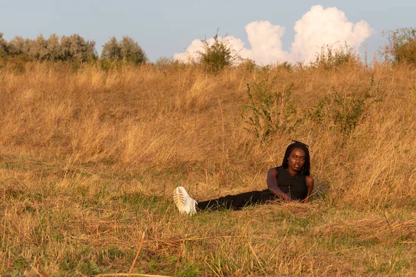 Chica africana en ropa negra se sienta entre el campo y disfruta de un día soleado de verano — Foto de Stock