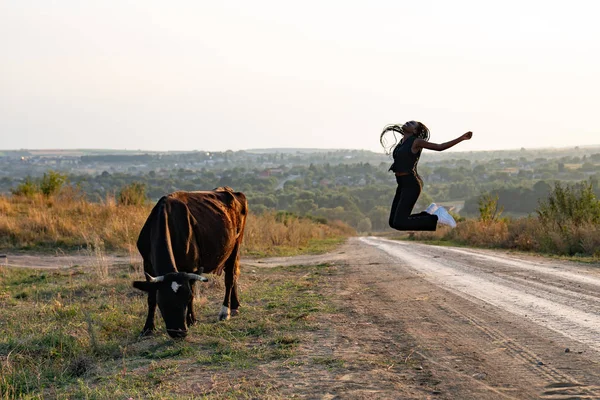 검은 옷을 입은 행복 한 아프리카 소녀가 비포장 도로에서 뛰고 소가 소녀 근처에서 풀을 뜯고 있는 모습 — 스톡 사진