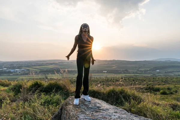 Mutlu gülümseyen siyah elbiseli Afrikalı kız büyük taşın üzerinde duruyor elleri açık, arka planda gün batımı — Stok fotoğraf