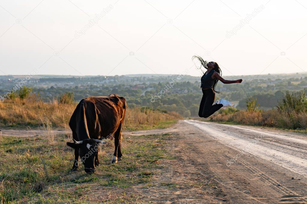 Happy african girl in black clothes jump on a dirt road, cow graze near the girl