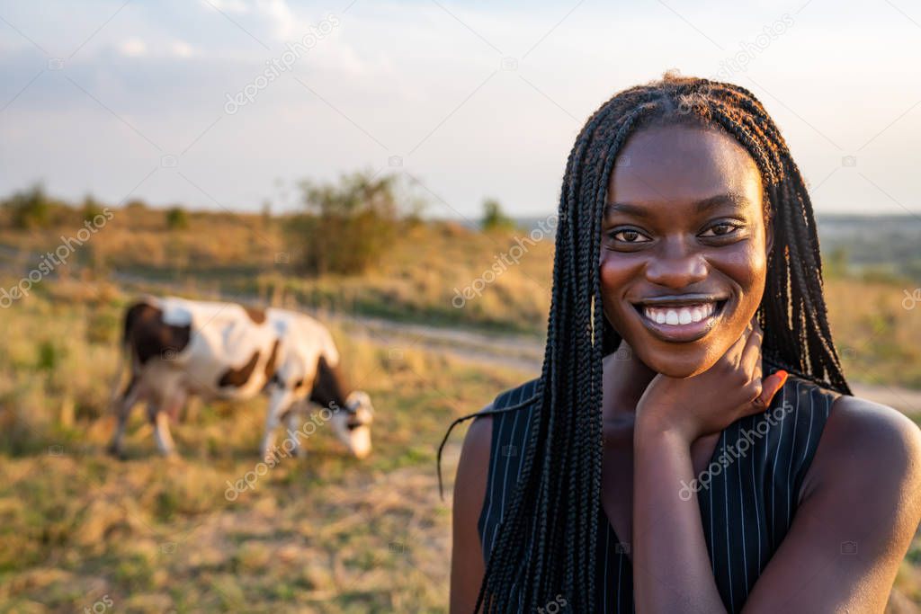 Close up portrait of the young african girl in black vest among the field, cow graze on the background