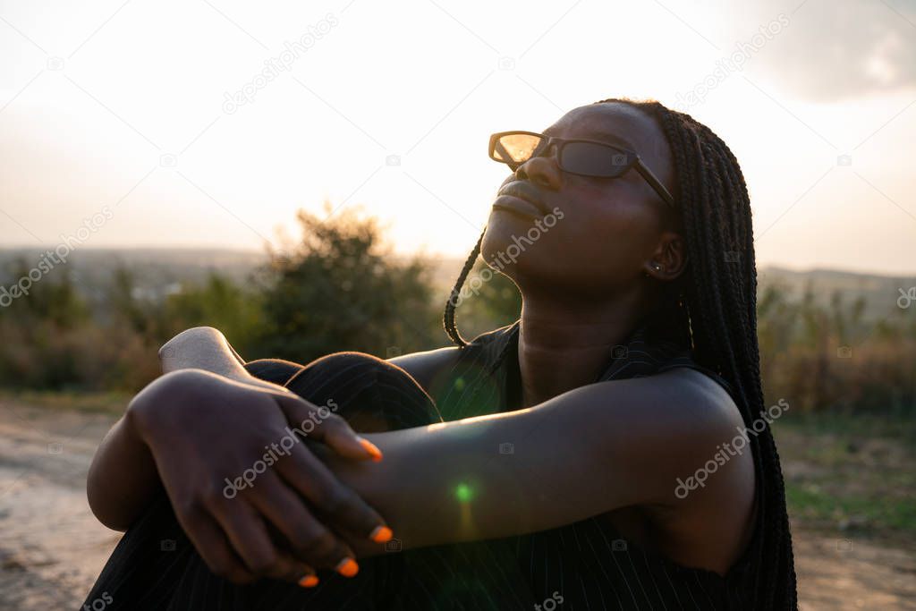 Young african girl in black sunglasses sits on the dirt road, hugging her knees and looking up, sunset on the background