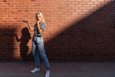 Gri tişörtlü ve mavi kotlu düşünceli sarışın kız kırmızı tuğla duvarın üzerinde, açık avuç içinizi tutuyor.