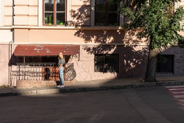 Young teen blonde girl in casual clothes stands in the sun on the sidewalk near the old building among the old city center