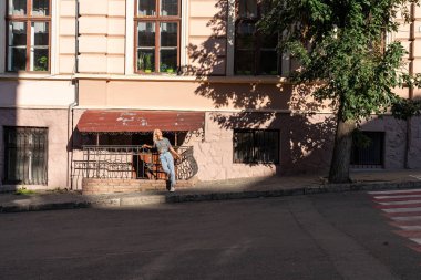 Young teen blonde girl in casual clothes stands in the sun on the sidewalk near the old building among the old city center