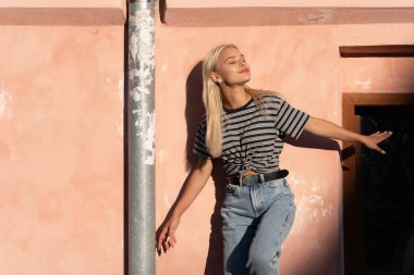 Çekici, rüya gibi, sarı saçlı, gri tişörtlü kız. Güneşte duruyor, eski pembe duvarın üzerinde.