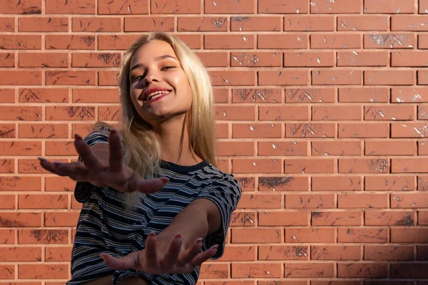 Glad leende blond flicka i grå randig t-shirt drar in händerna i kameran isolerad över röd tegelvägg — Stockfoto