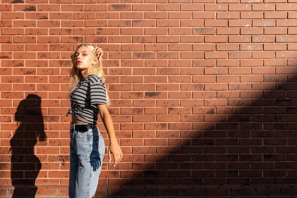 Attractive blonde teen girl in grey striped y-shirt and jeans touching her hair and lookinf at the camera isolated over red brick wall — Stock Photo, Image