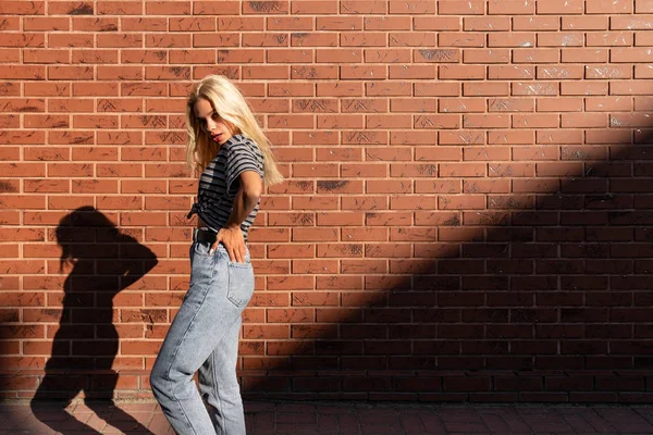 Visão lateral retrato de jovem loira em gey t-shirt e jeans que syands no sol, segura as mãos em sua cintura, isolado sobre parede de tijolo vermelho — Fotografia de Stock