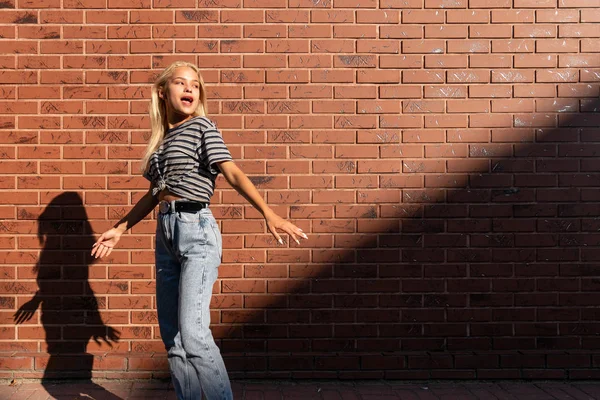 Atractiva chica adolescente rubia en camiseta gris y jeans bailando al sol aislados sobre la pared de ladrillo rojo — Foto de Stock