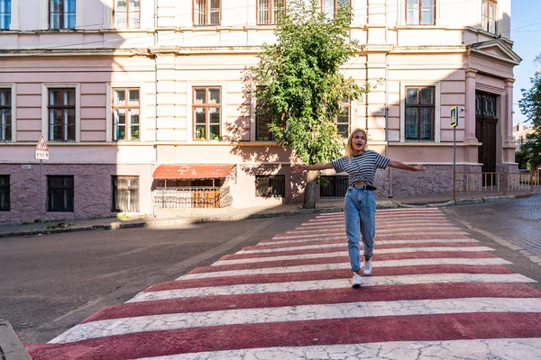 Estate ritratto soleggiato della giovane attraente viaggiatrice bionda in t-shirt grigia e jeans blu sulla passerella rossa tra il centro storico della città — Foto Stock