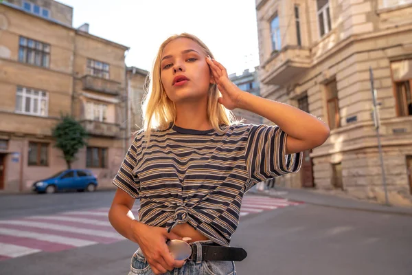 Περιμένετε μέχρι το πορτρέτο του νεαρού έφηβος ξανθιά κοπέλα με χαλαρά μαλλιά ντυμένος με γκρι ριγέ t-shirt βόλτες μεταξύ του κέντρου της παλιάς πόλης, ηλιοβασίλεμα στο παρασκήνιο — Φωτογραφία Αρχείου