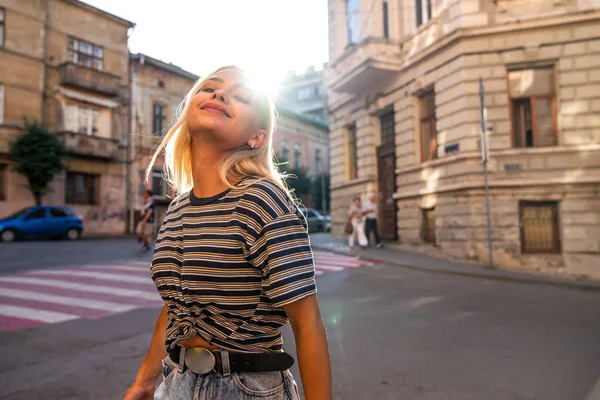 Талия до портрета молодой девушки-блондинки с распущенными волосами, одетые в серые полосатые футболки прогулки среди старого центра города, закат на заднем плане — стоковое фото