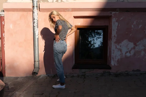 Chica rubia de moda con el pelo suelto vestida con camiseta gris se pone al sol cerca de la pared rosa de la antigua casa y toma las manos en la cintura — Foto de Stock