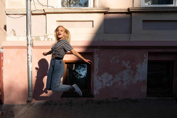 Feliz excitada chica adolescente rubia con el pelo suelto vestido con ropa casual saltando entre la calle de la ciudad vieja — Foto de Stock