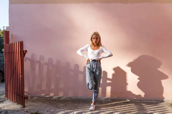 Chica rubia de moda en la parte superior blanca y pantalones vaqueros azules se para en una pierna al sol y mirando a la cámara, la pared rosa en el fondo — Foto de Stock
