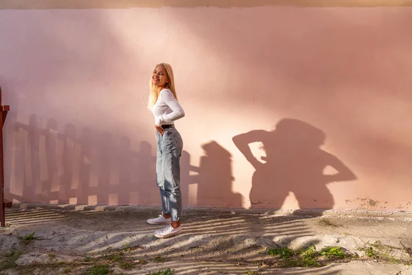 Feliz sonriente rubia adolescente chica en casual clotehs sostiene sus manos en su cintura y mirando a la cámara, rosa pared en el fondo — Foto de Stock