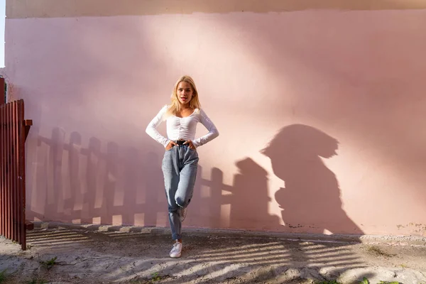 Menina loira na moda em top branco e jeans azul fica em uma perna ao sol e olhando para a câmera, parede rosa no fundo — Fotografia de Stock