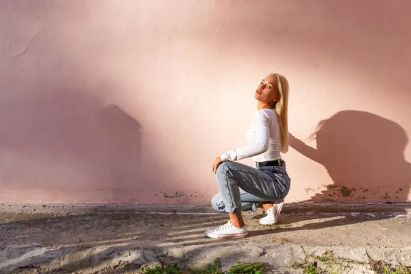 Atractiva chica rubia trandy en la parte superior blanca agachado cerca de la pared rosa y mirando a la cámara — Foto de Stock