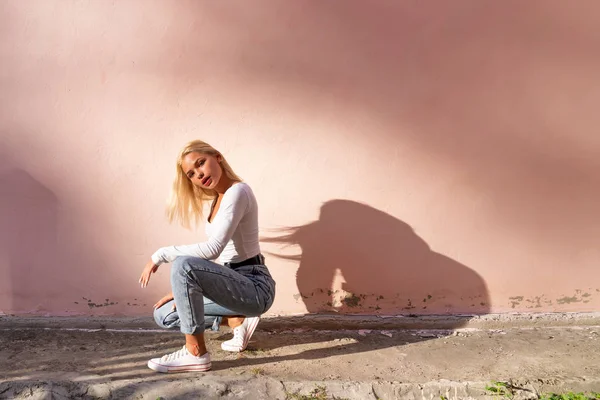 Atrakcyjne trandy blondynka dziewczyna w biały top przykucnął w pobliżu różowej ściany i patrząc w aparat — Zdjęcie stockowe