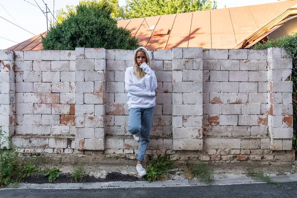 Joven adolescente en sudadera con capucha blanca apoyada en la pared de ladrillo blanco y mirando a la cámara — Foto de Stock