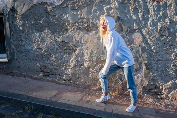 Street style fashion portrait of the blonde trendy girl in white hoodie near the old grey wall