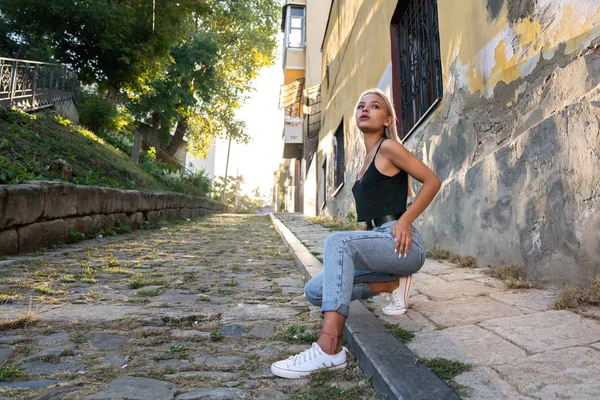 Retrato al estilo de la calle de la hermosa chica rubia en ropa casual que se agachó entre las piedras de la calle, puesta de sol en el fondo — Foto de Stock