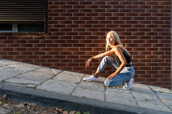 Chica rubia adolescente de moda en ropa casual agachada cerca de la pared de ladrillo rojo, los rayos del sol están cayendo en su cara — Foto de Stock