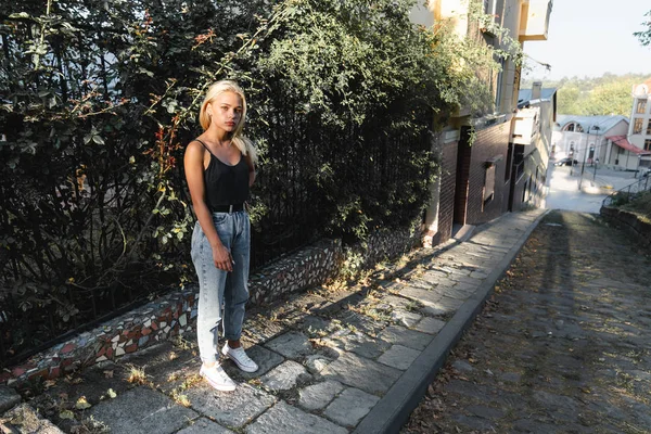 Attarctive tranquila chica rubia en ropa casual se encuentra cerca de la cerca verde entre la vieja calle de la ciudad — Foto de Stock