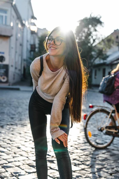 Pretty cute brunette girl traveler in casual clothes with black backpack stands among the paving stones street, enjoying sunny weekend in the old european city
