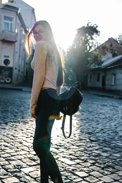 Linda chica morena viajera en ropa casual con mochila negra se encuentra entre las piedras de la calle, disfrutando de un soleado fin de semana en la vieja ciudad europea — Foto de Stock