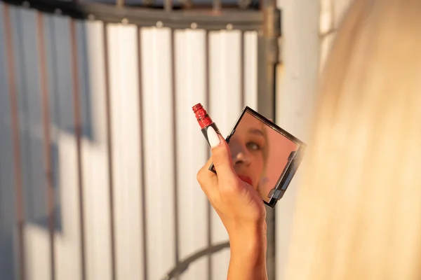 Blondes Mädchen hält roten Lippenstift in der Hand und blickt durch den Spiegel in die Kamera — Stockfoto