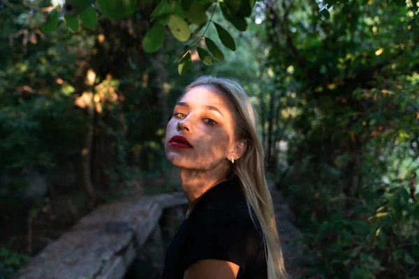 Close up retrato da linda menina loira com lábios vermelhos e pele saudável, fica entre os arbustos verdes na sombra — Fotografia de Stock