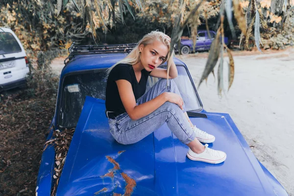 Street style fashion portrait of the blonde girl in black top and blue jeans that sits on the brocken blue old retro car and looking at the camera — Stock Photo, Image