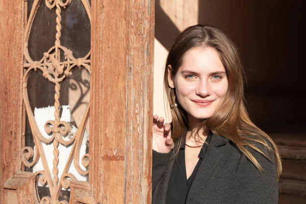 Fille brune à la mode avec un maquillage naturel, vêtue d'un manteau noir tient sa main sur la porte en bois vintage et regarde la caméra — Photo