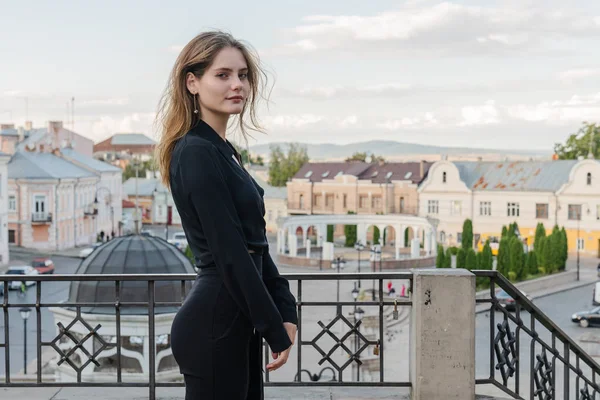 Hermosa chica elegante con morena suelta cabello ondulado, vestida con camisa negra y pantalones se para en el balcón y mirando a la cámara, ciudad vieja en el fondo — Foto de Stock