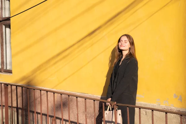 Menina bonita em preto cloack e bolsa em mãos apoiadas na parede amarela e desfrutar do sol com os olhos fechados — Fotografia de Stock