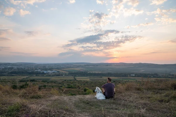 Jeune homme assis avec son chien samoyed parmi les champs et profiter d'un paysage incroyable avec coucher de soleil — Photo