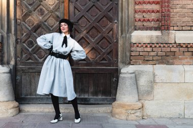 Eski moda elbiseli ve siyah bereli mutlu genç bayan ellerini belinde tutuyor ve kameraya bakıyor. Arka planda eski, ahşap bir kapı var.