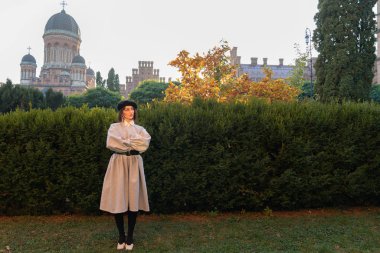 Siyah bereli ve gri eski moda elbiseli genç kadının tam boy portresi. Arka planda yeşil çalılar var.