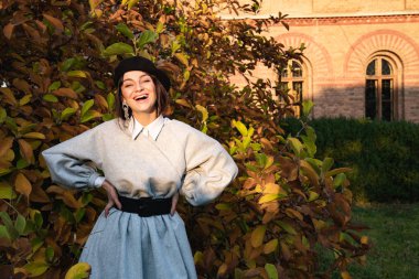 Siyah bereli ve gri elbiseli mutlu kız sonbahar ağacının yanında duruyor. Arka planda eski tuğla bina.