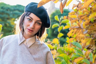 Siyah bereli ve gri elbiseli çekici genç bir kadın sonbahar çalılarının arasında dikilip kameraya bakıyor.