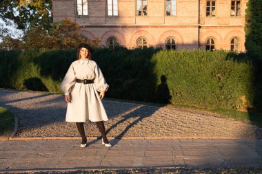 Siyah bereli ve gri eski moda elbiseli çekici genç bir kadın eski tuğla binanın yanındaki güneşte duruyor.