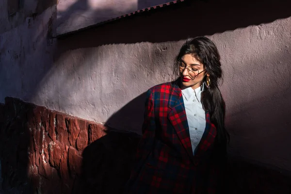 Menina morena atraente em óculos, vestida com jaqueta xadrez e camisa branca apoiada na parede e olhando para o canto esquerdo para baixo, raios de sol caem em seu rosto — Fotografia de Stock